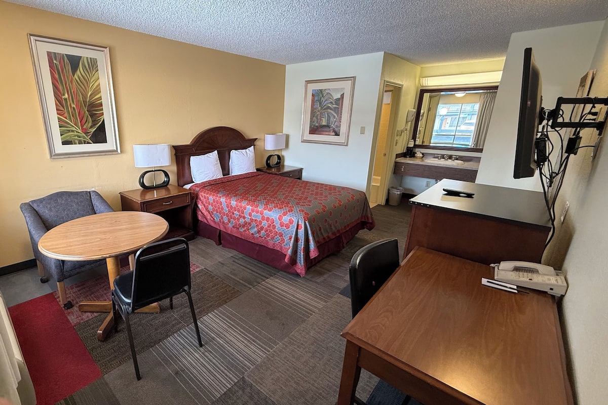 A hotel room with a bed, a desk, a chair, a small table, and a lamp.
