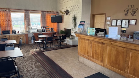 A hotel lobby with a reception desk, a dining area with tables and chairs, a kitchen area, and a large window with curtains.