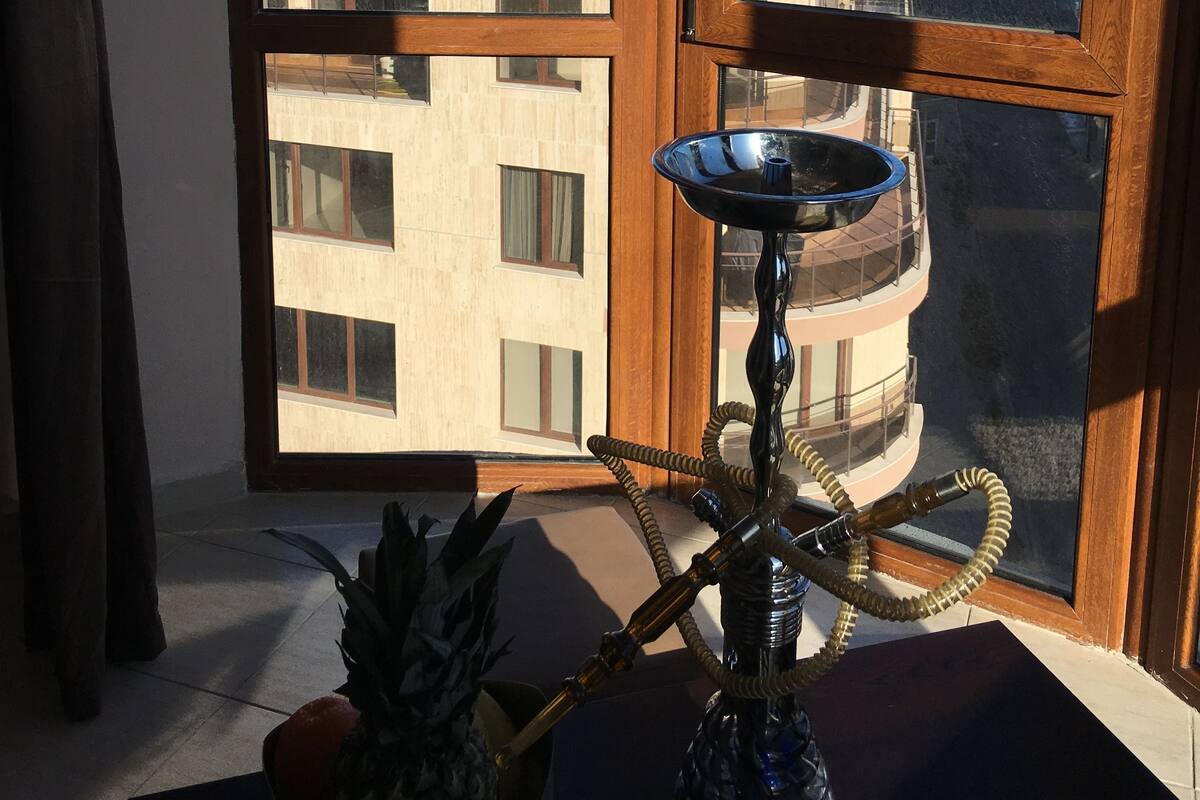 A room with a view of the sea through a large window, a hookah on a table, and a pineapple on the windowsill.