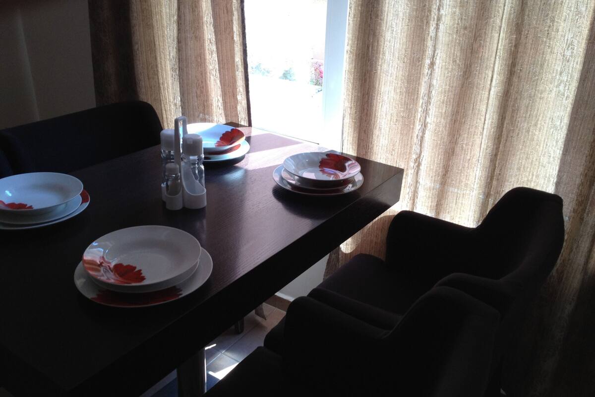 A dining table with plates and cutlery, set against a window with curtains.