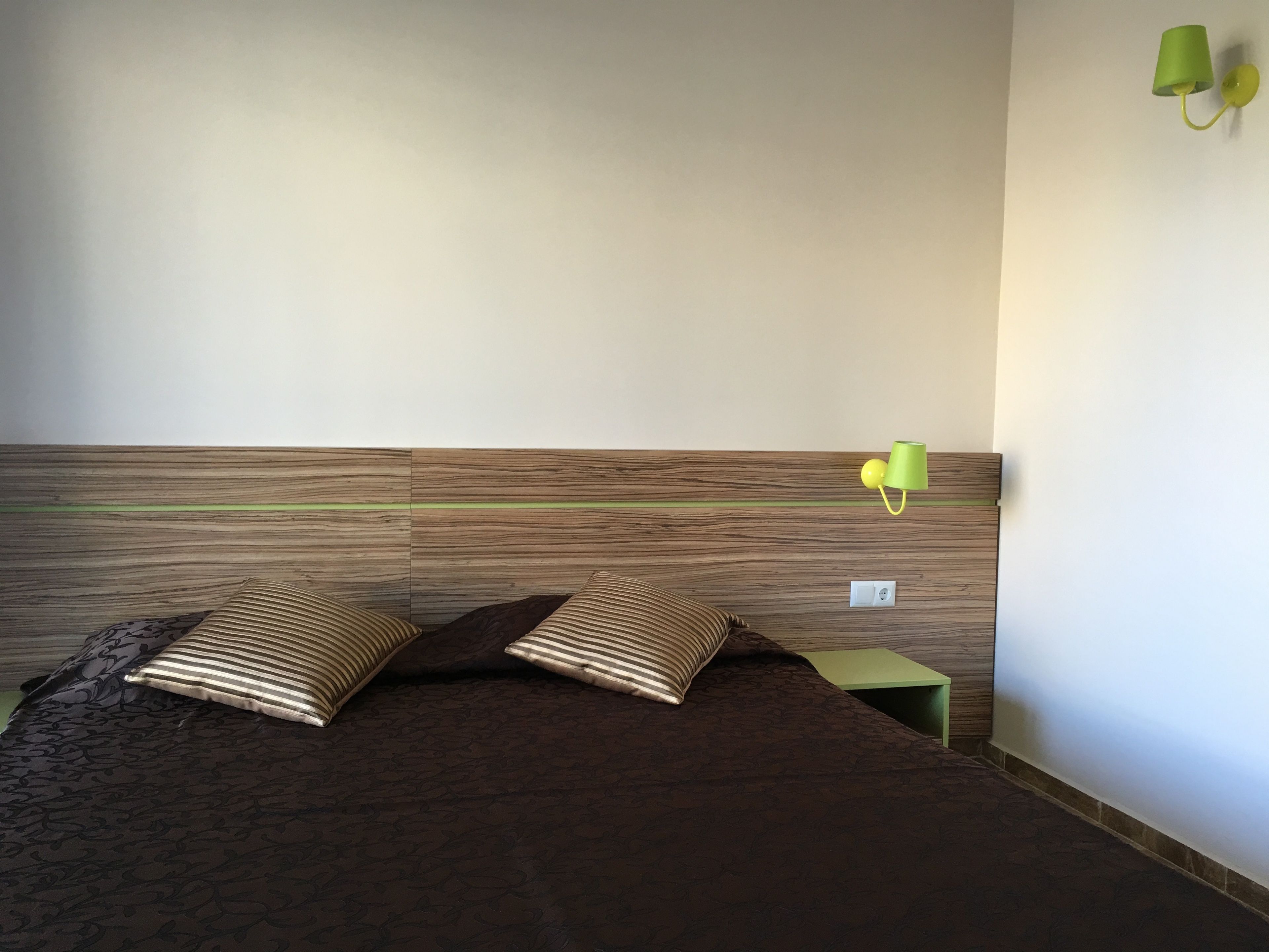 A bedroom with a wooden headboard, two striped pillows, a green bedside table, and a green wall lamp.