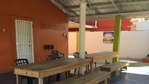 Un área de estar al aire libre cubierta con una mesa de madera larga y bancos, rodeada de paredes anaranjadas y un piso de baldosas.