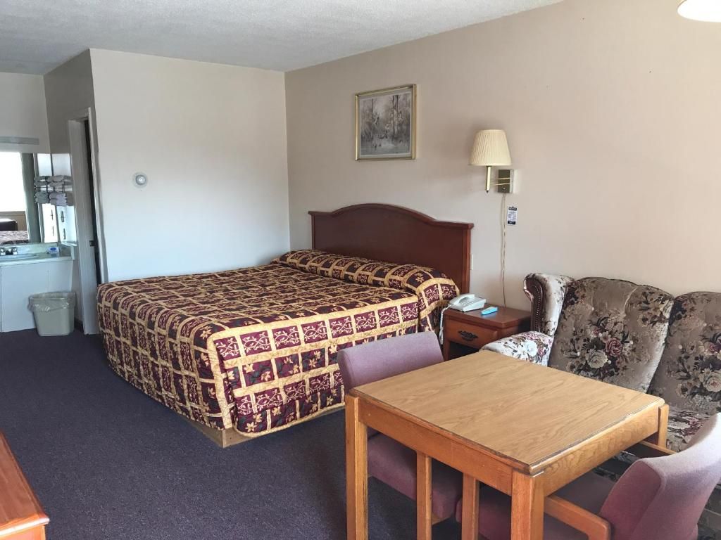 A hotel room with a bed, a table, a chair, a sofa, a lamp, and a painting on the wall.
