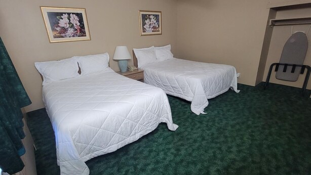 A hotel room with two single beds, a green carpet, a small table with a lamp, and two framed floral pictures on the wall. - Medina Inn (Medina)