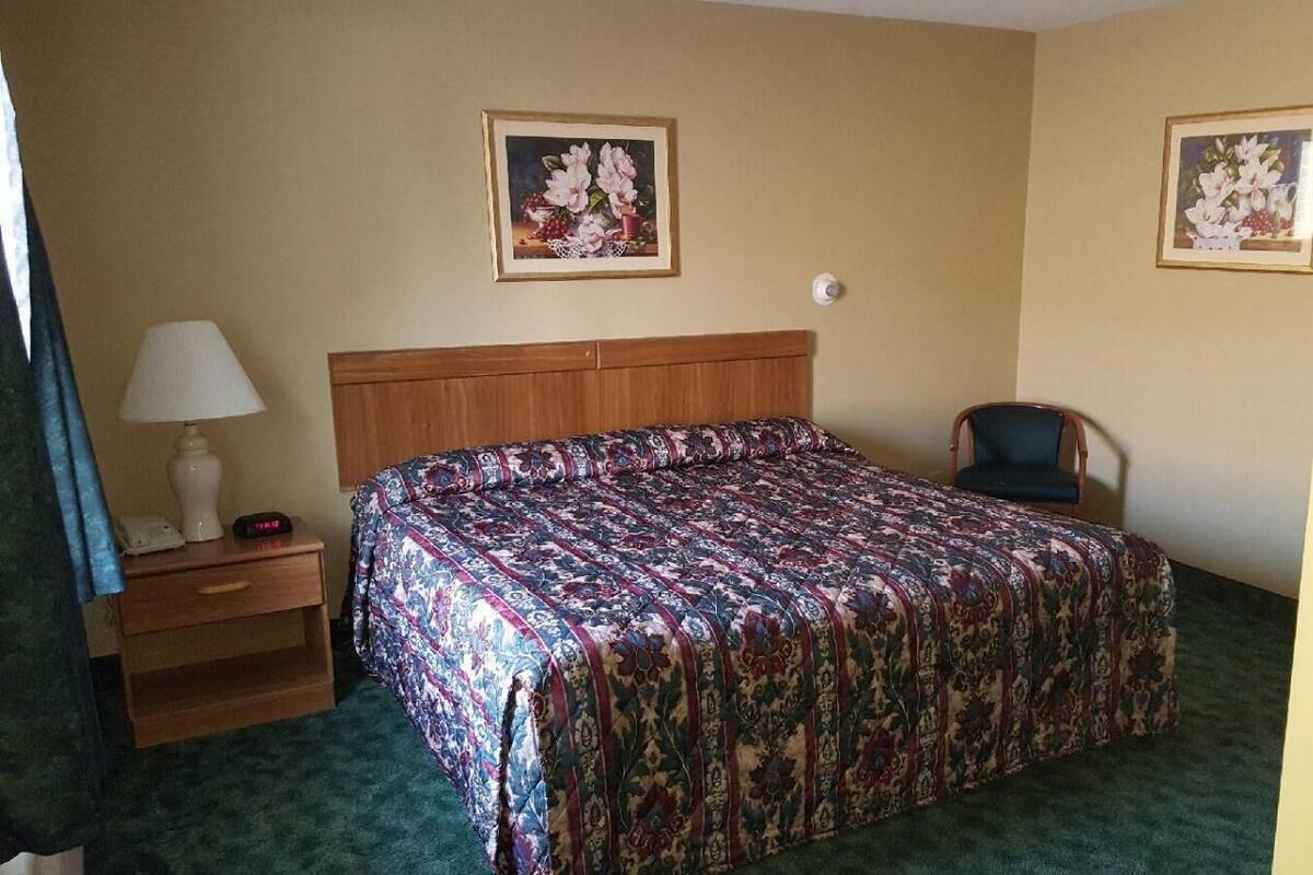 A hotel room with a bed, a chair, a nightstand with a lamp, and two framed floral paintings on the walls.