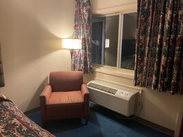 Quarto de hotel com uma cadeira, janela com cortinas, ar condicionado fixado na parede e uma luminária.