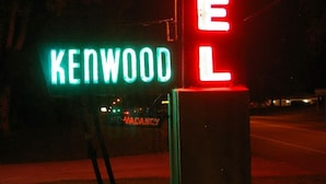 Un letrero de motel neón con el nombre 'KENWOOD' y 'MOTEL' en luces rojas y verdes brillantes, respectivamente, montado sobre un poste de concreto.
