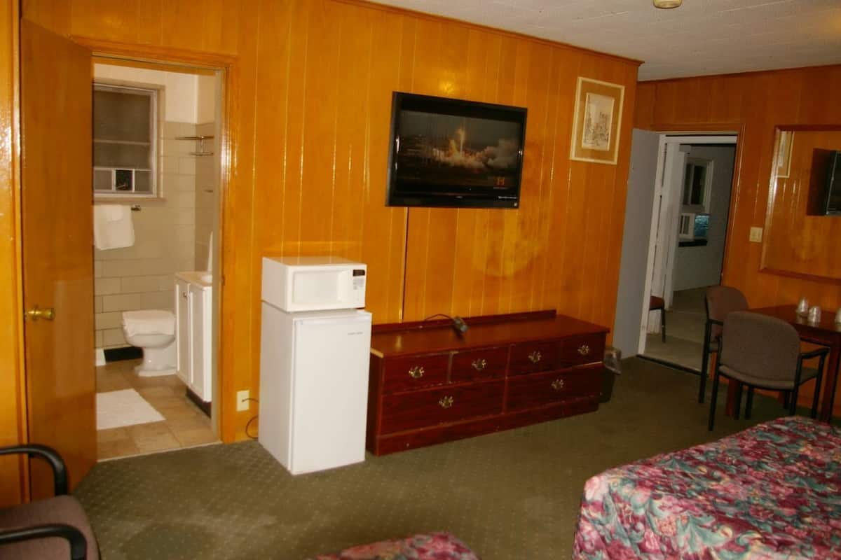 A hotel room with wooden paneling, a television mounted on the wall, a small refrigerator, a wooden dresser, a bed with a floral bedspread, and a bathroom with a toilet and sink.