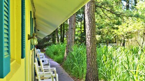 A covered outdoor seating area with white chairs and a wooden deck, surrounded by tall trees and lush greenery. - Meris Gardens Bethany (Bethany Beach)