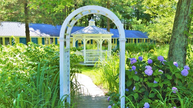 A wooden pathway leading to a white gazebo with a blue roof, surrounded by lush greenery and purple flowers. - Meris Gardens Bethany (Bethany Beach)