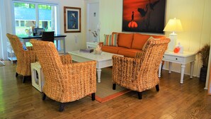 A living room with wicker furniture, a white coffee table, an orange sofa, and a painting of birds. - Meris Gardens Bethany (Bethany Beach)