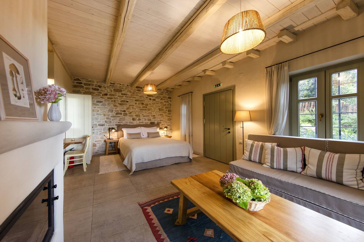 A cozy bedroom with a stone wall, a wooden ceiling, a bed, a bench with cushions, and a table with a vase of flowers.