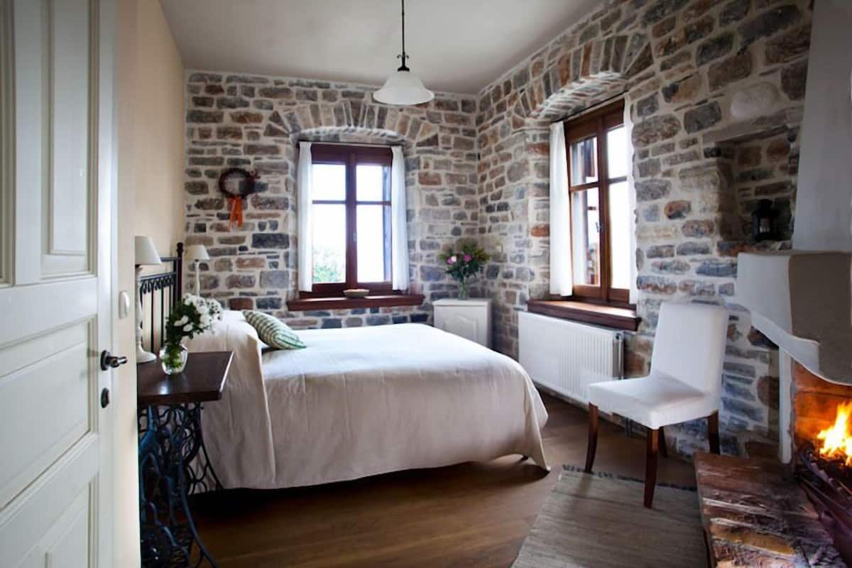 A bedroom with a stone wall, a fireplace, a bed, a chair, and a window with curtains.