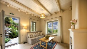 A room with a wooden table, a sofa, a lamp, and a window with a view of greenery. - Amanita Guesthouse (Zagora-Mouresi)