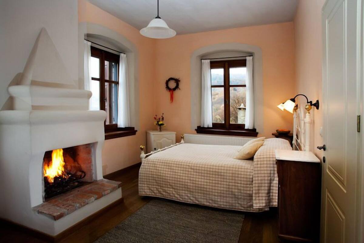A bedroom with a fireplace, a bed with a checkered bedspread, two windows with curtains, and a wall-mounted light fixture.
