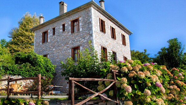 A stone building with multiple windows and a chimney, surrounded by lush greenery and flowering plants. - Amanita Guesthouse (Zagora-Mouresi)
