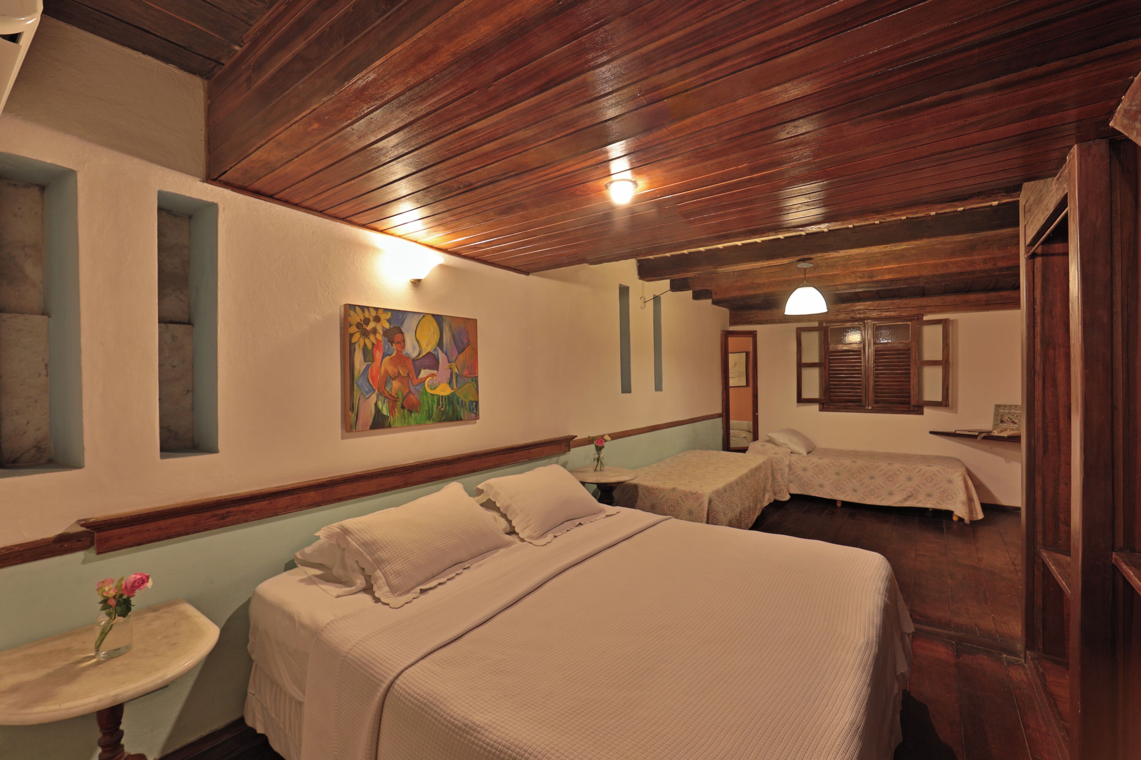 A room with a wooden ceiling, a bed with a white bedspread, a small table with a vase of flowers, and a painting on the wall.