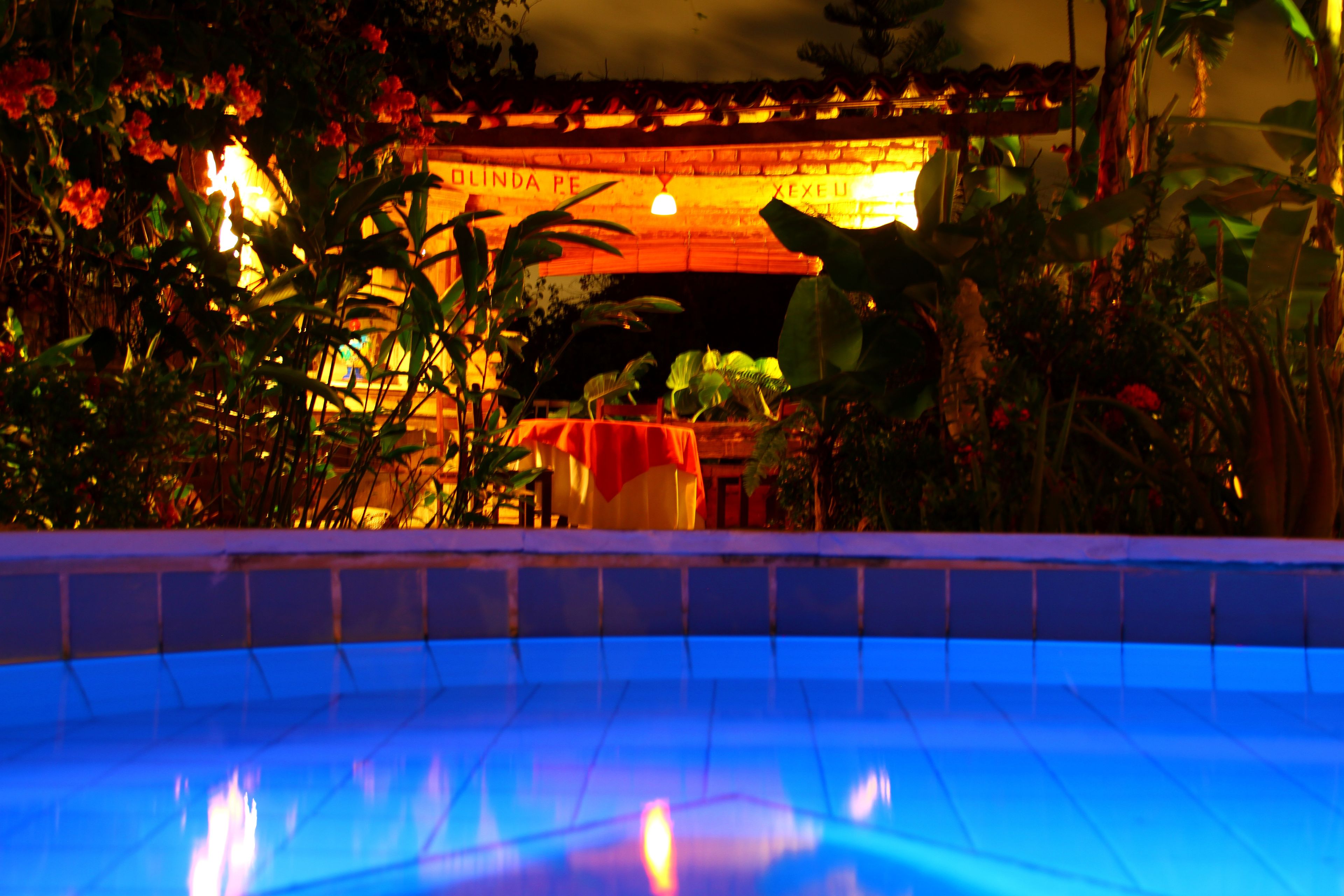 Una piscina con borde de azulejos, rodeada de abundante vegetación y un edificio con un cartel que dice 'OLINDA PE'.