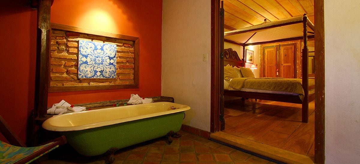 A bathroom with a green clawfoot bathtub, a wooden door leading to a bedroom, and a stone wall with a blue and white tile pattern.