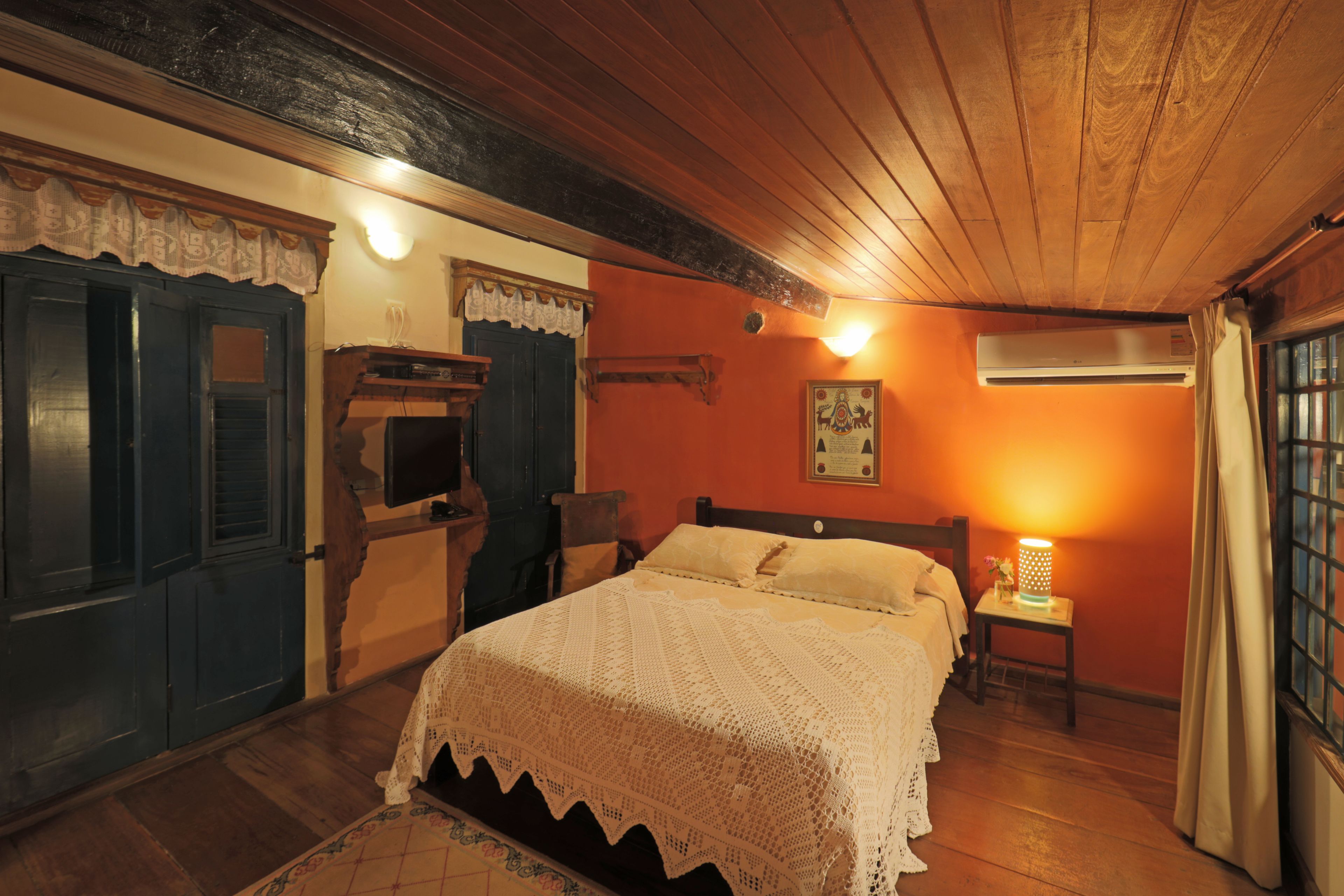 A cozy bedroom with a wooden ceiling, a bed with a white blanket, a nightstand with a lamp, and a window with curtains.