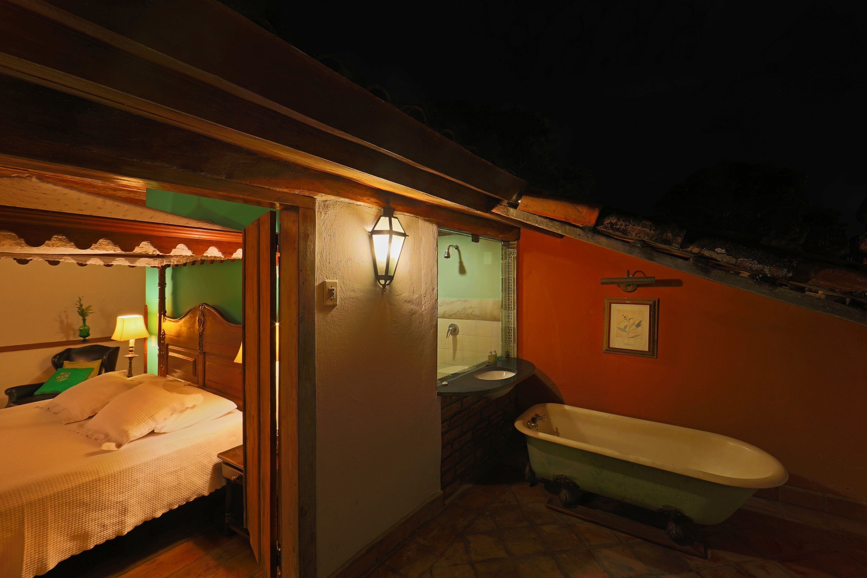 Una habitación pequeña con luz tenue, que incluye una cama, un mesita de noche, una lámpara y un baño con bañera y ducha.