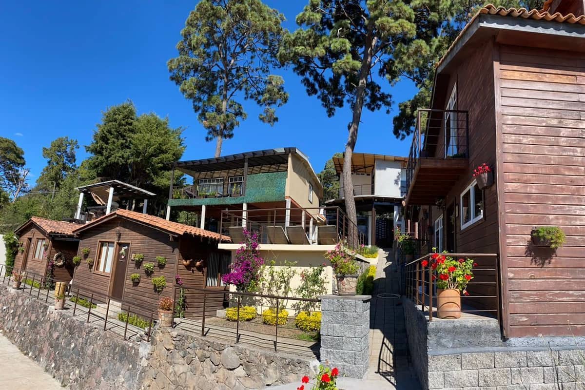 Una hilera de cabañas de madera con balcones, rodeadas de muros de piedra y diversas plantas.