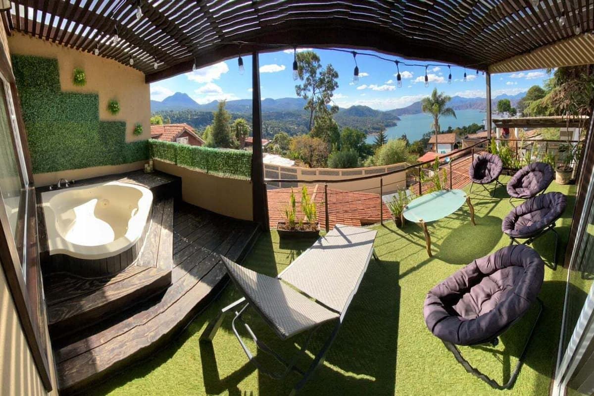 Un espacio exterior techado con un jacuzzi, asientos y vistas a montañas y un lago.