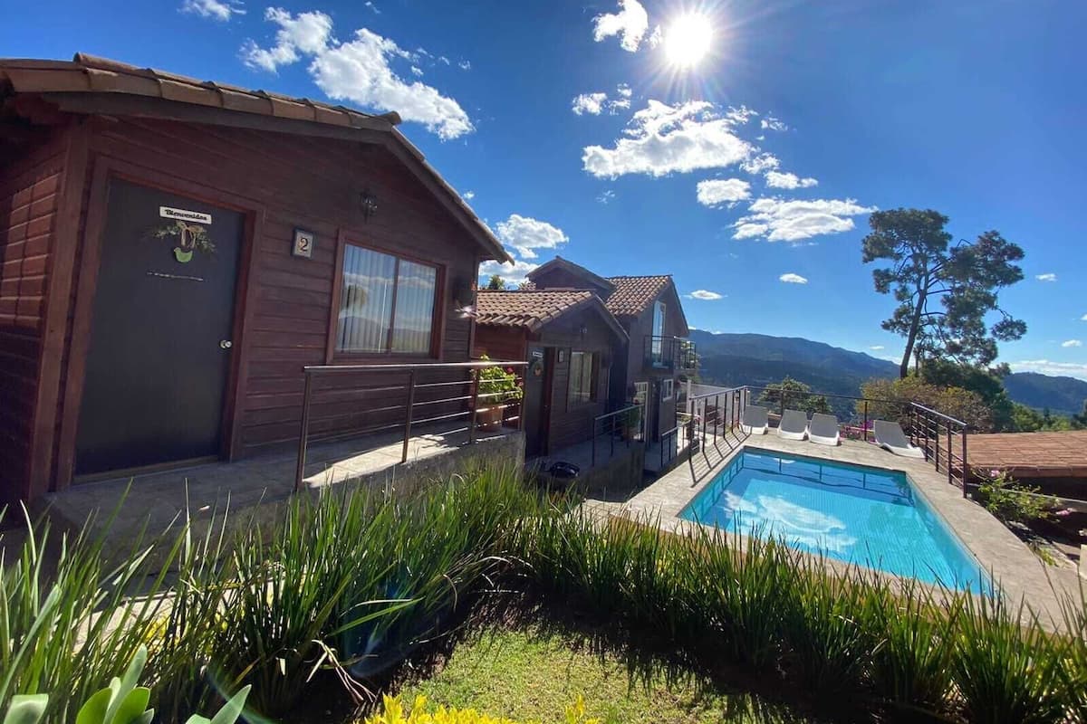 Una cabaña de madera con piscina, rodeada de vegetación y montañas al fondo.