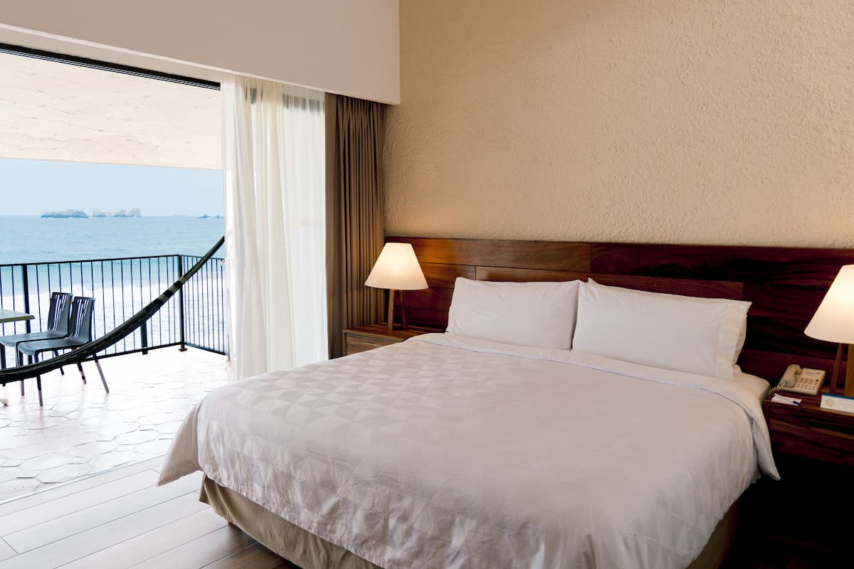 A hotel room with a large bed, a nightstand with a lamp, a chair, and a view of the sea.