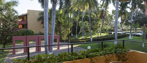 A balcony with a view of a landscaped garden, palm trees, and a building with a red structure.