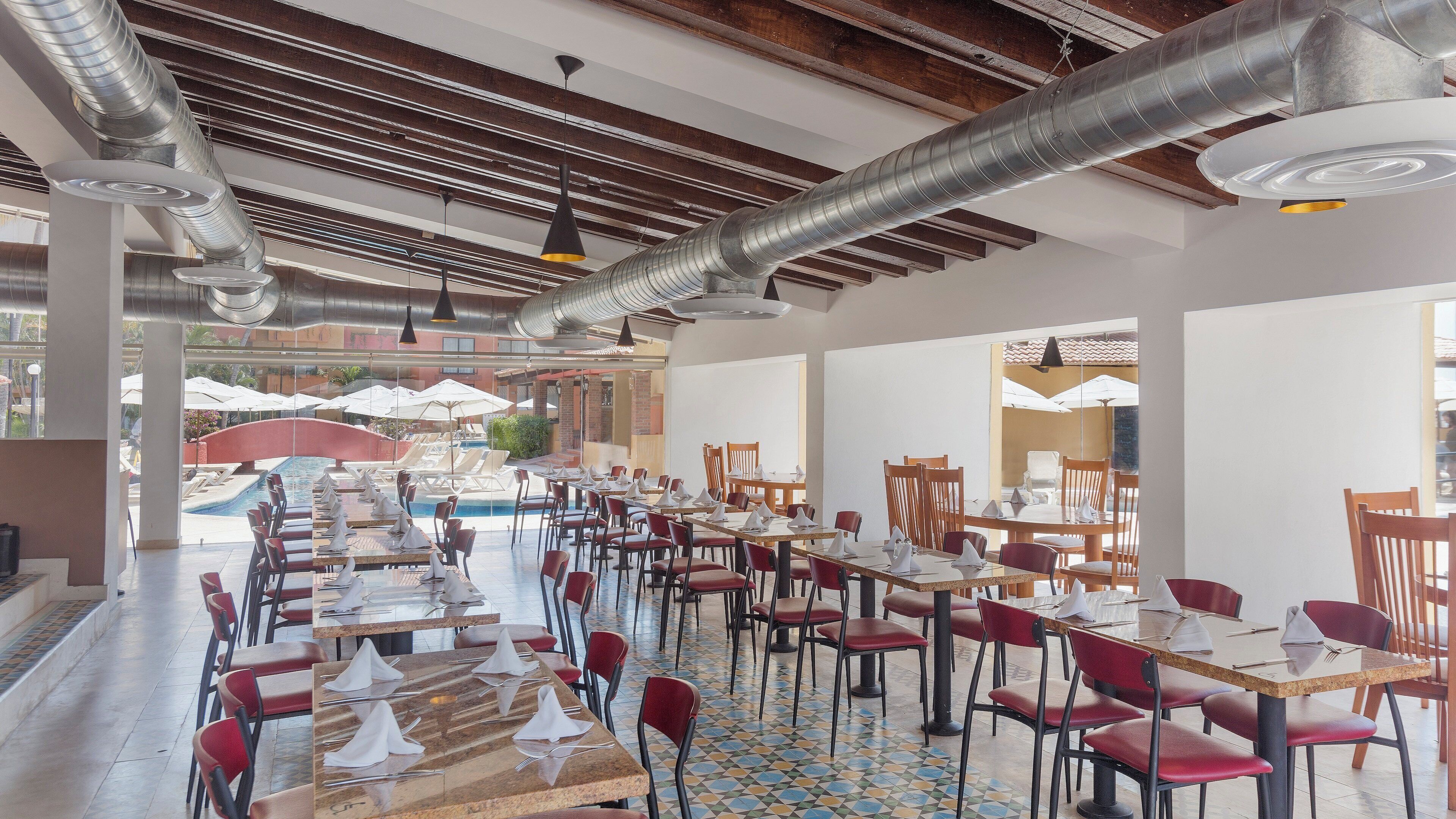 Un vaste espace dédié à la salle à manger, avec de nombreuses tables et chaises, une piscine en arrière-plan et un plafond moderne aux poutres apparentes et aux conduits visibles.