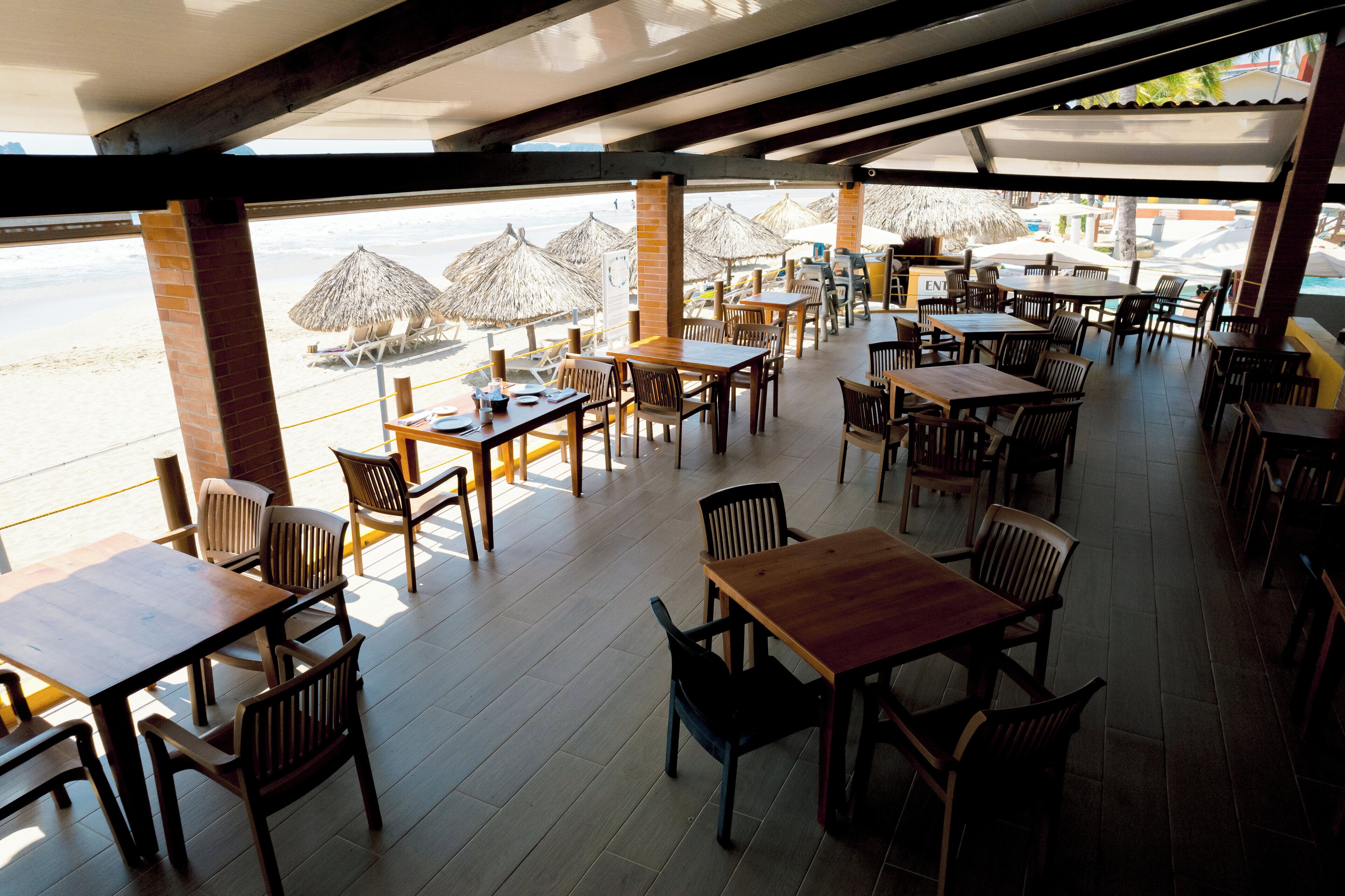Un espacio de comedor al aire libre con mesas y sillas de madera, sombrillas y vistas a la playa.