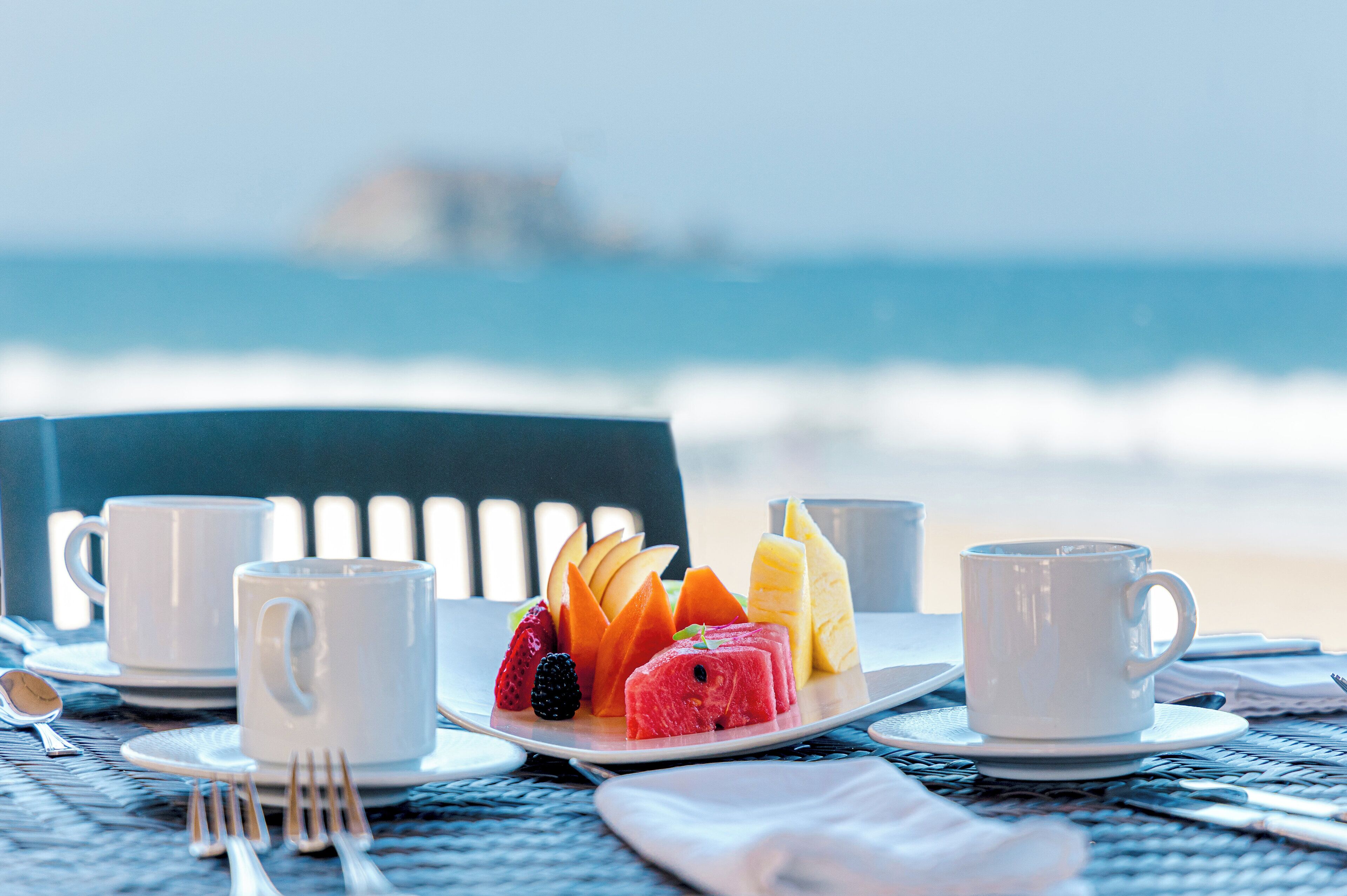 Mesa posta com xícaras brancas, prato de frutas e talheres numa praia, com uma cadeira ao fundo.