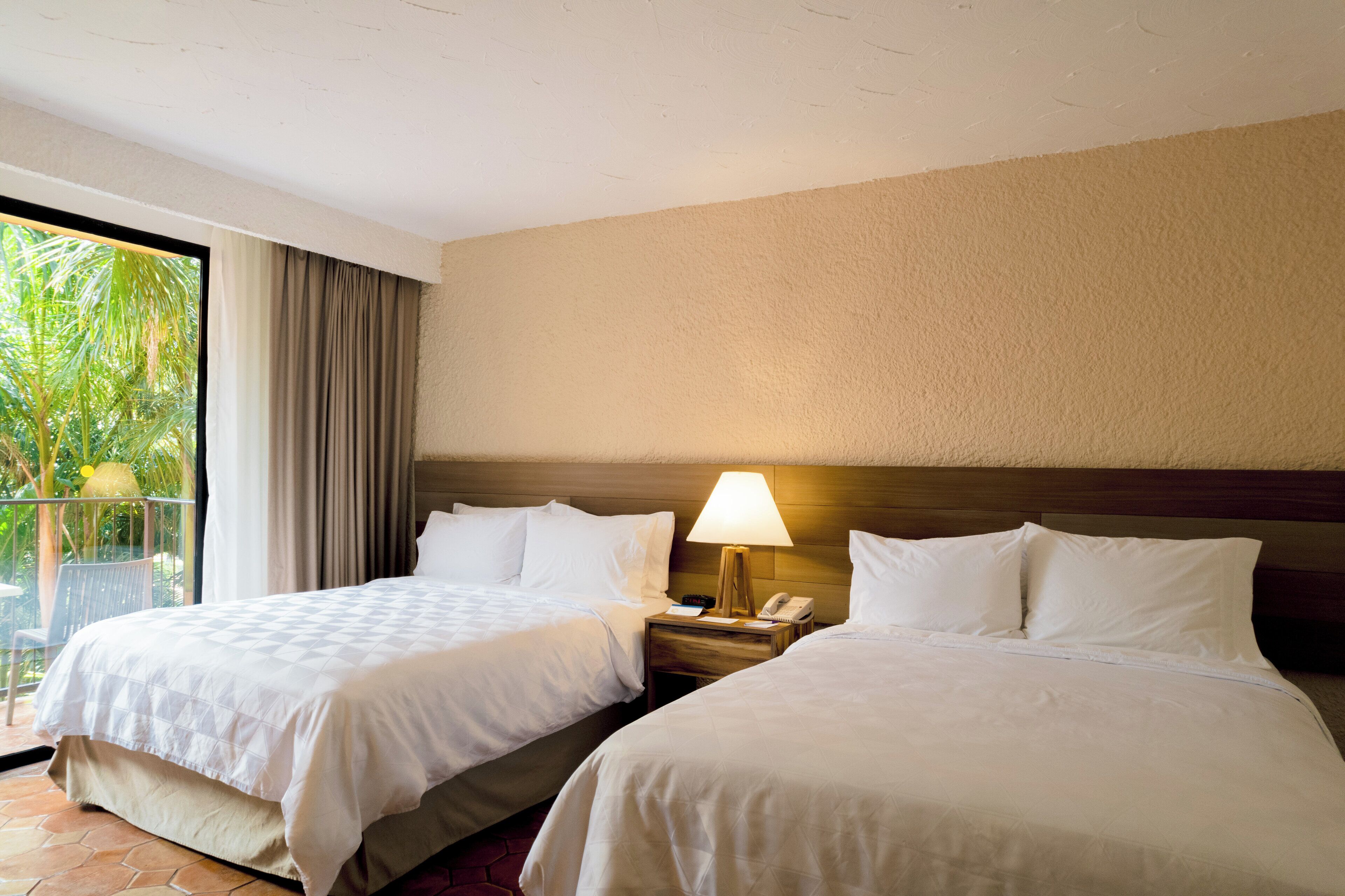 A hotel room with two beds, a nightstand with a lamp, and a balcony with a view of palm trees.