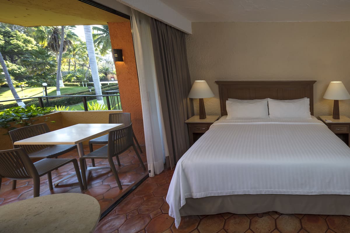 A hotel room with a bed, two bedside tables with lamps, a window with curtains, a small table with chairs, and a view of a garden with palm trees.