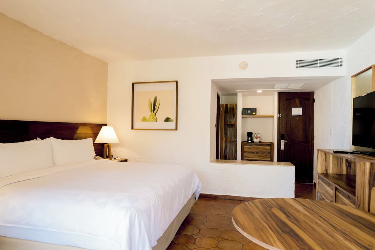 A hotel room with a large bed, a bedside table with a lamp, a wooden dresser, a television, and a framed picture on the wall.