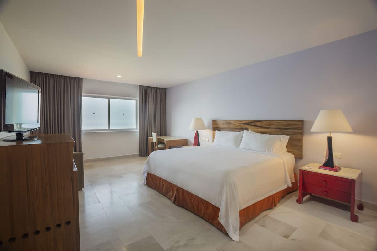 A hotel room with a large bed, two bedside tables with lamps, a desk, a TV, and a large window.