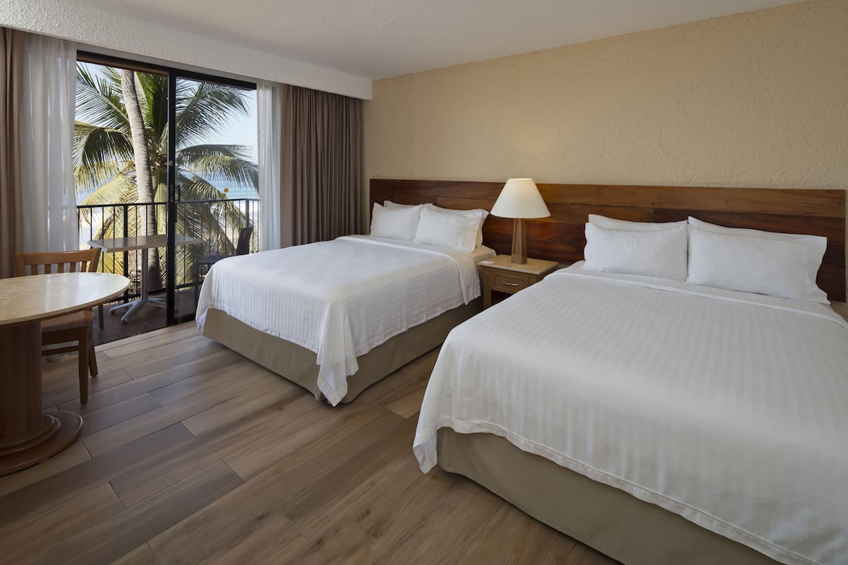 A hotel room with two beds, a wooden floor, a table, and a balcony with a view of palm trees.