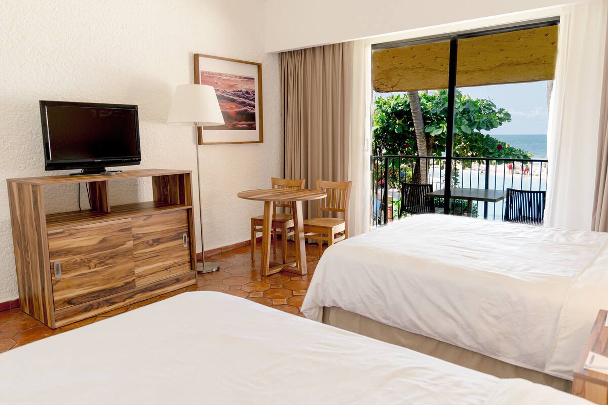 A hotel room with two beds, a TV, a desk, a chair, a table, and a balcony with a view of the beach.