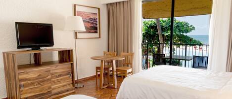 A hotel room with two beds, a TV, a desk, a chair, a table, and a balcony with a view of the beach.