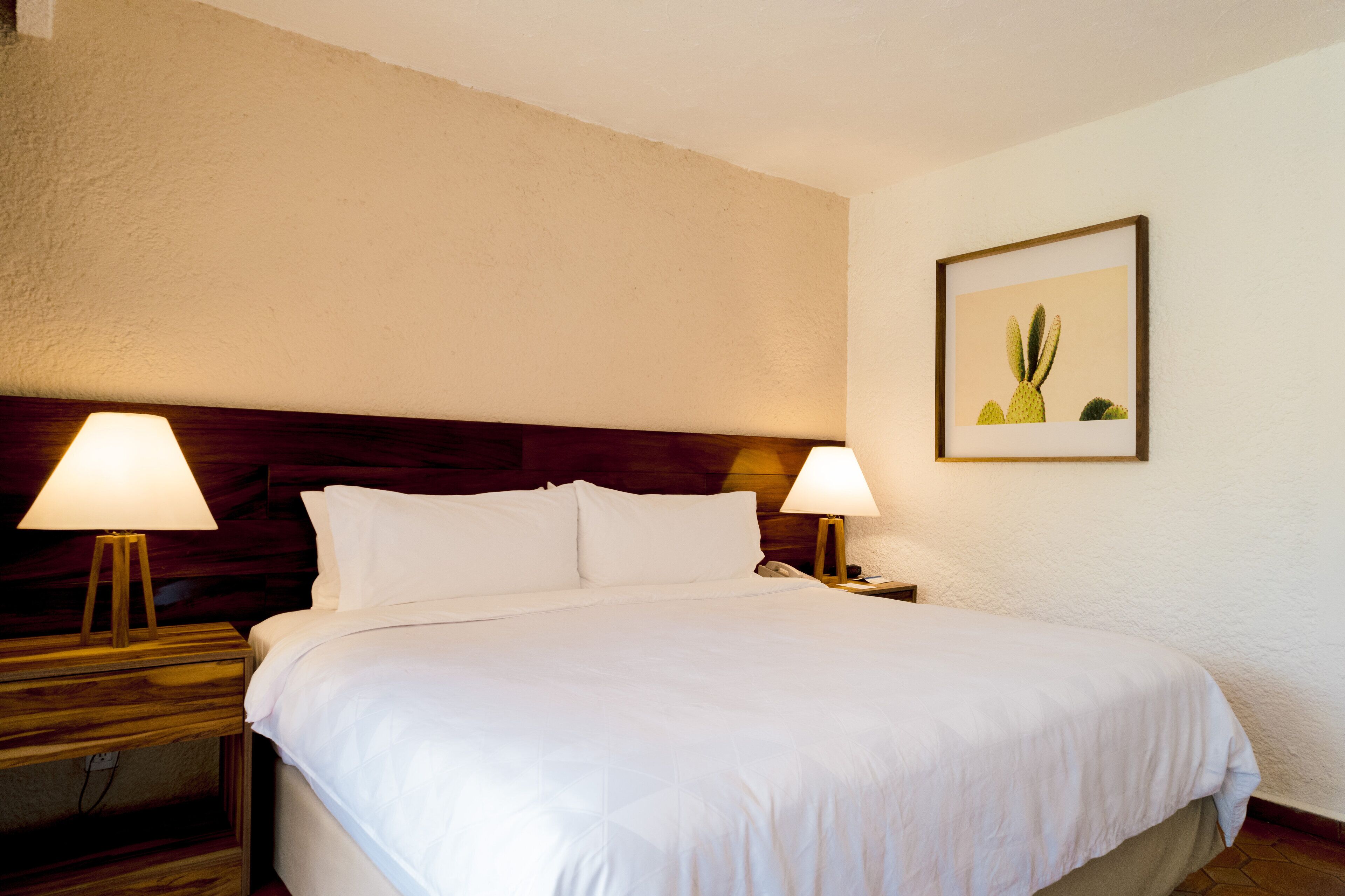 A neatly made bed with white linens, two bedside lamps, a wooden headboard, and a framed picture on the wall.