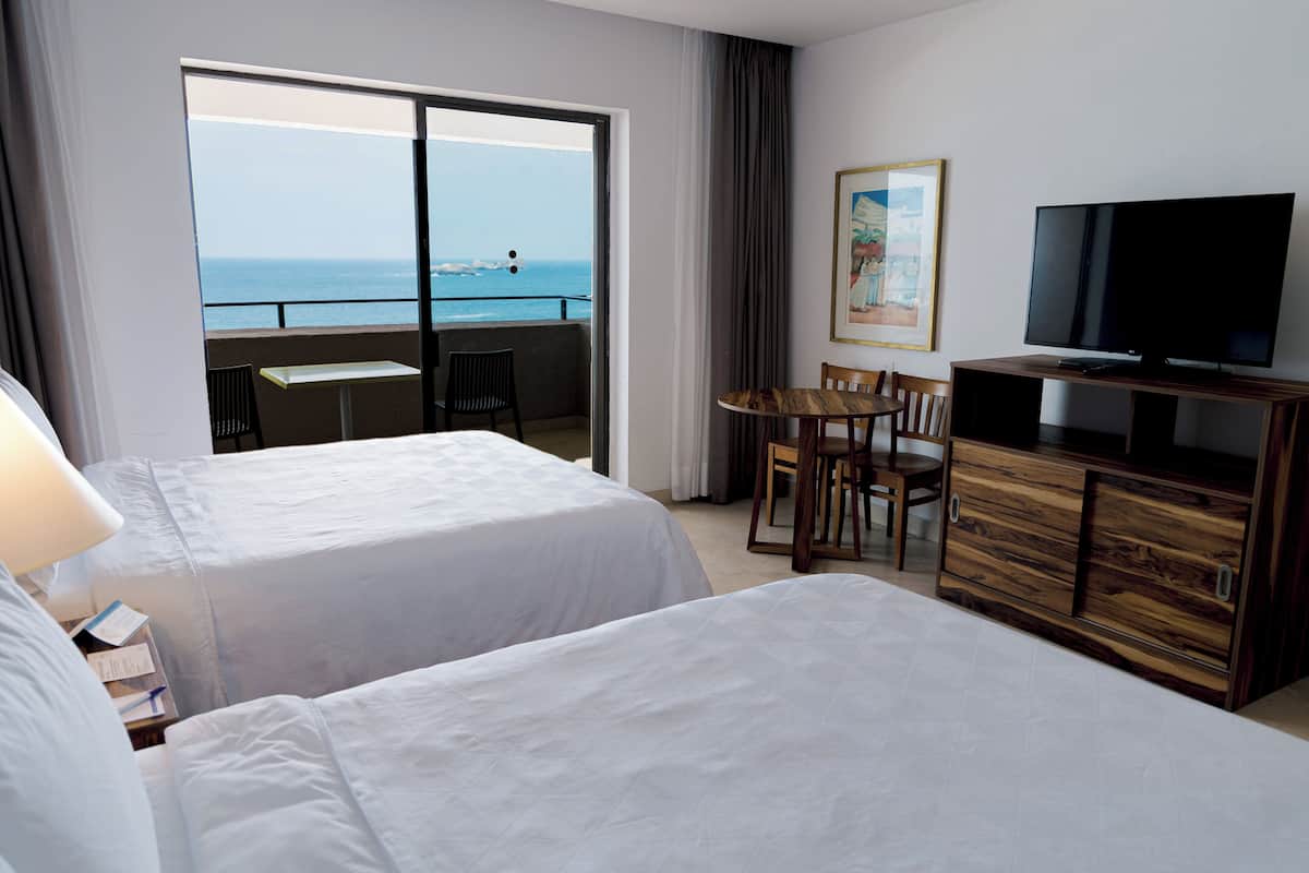 A hotel room with two beds, a TV, a dining table with chairs, and a view of the ocean.