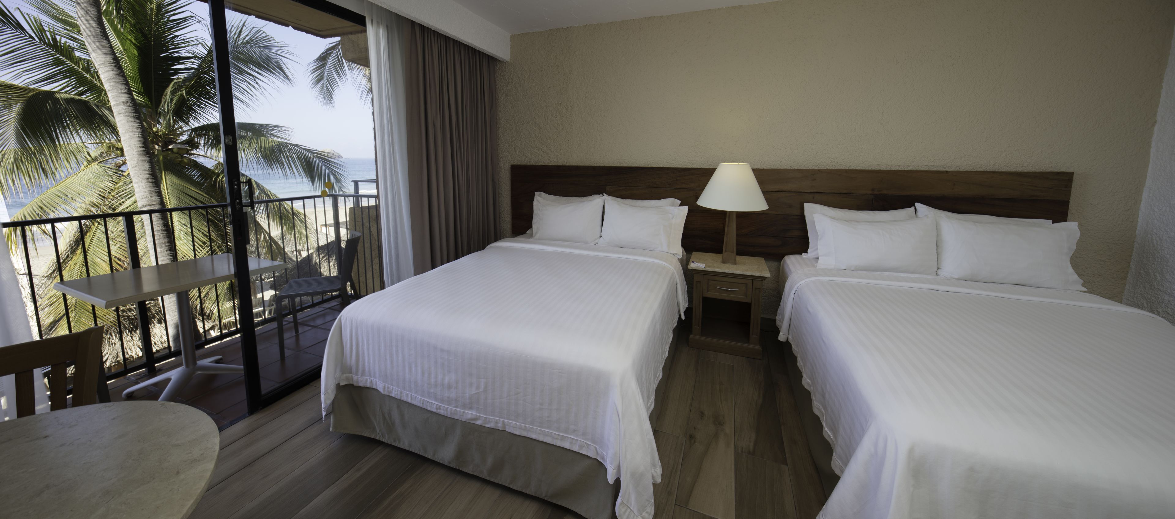 Habitación de hotel con dos camas, un balcón con mesa y sillas, y vista a palmeras y al océano.