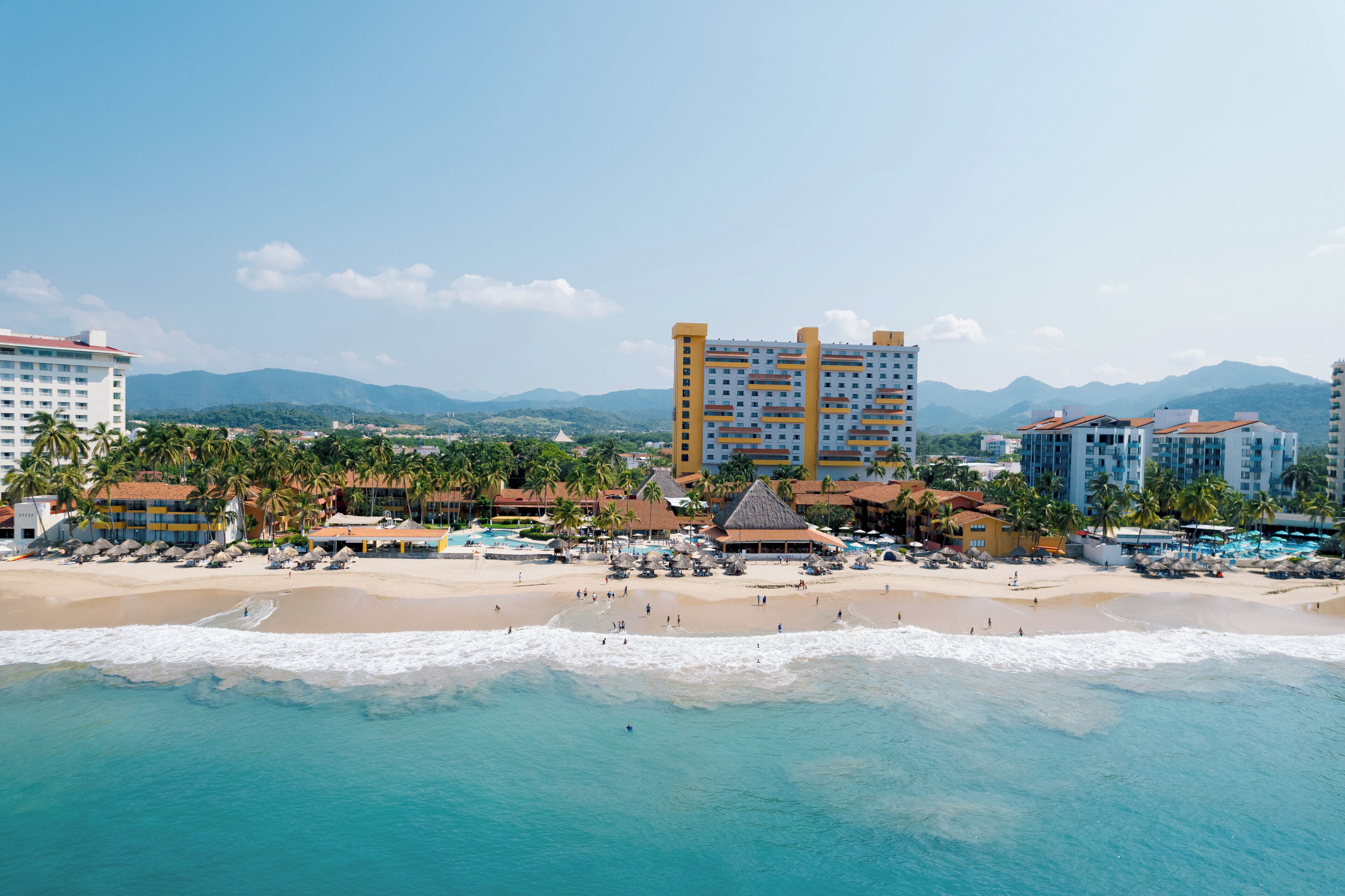 Un hotel frente a la playa con varias edificaciones, una playa de arena y una vista despejada del océano.