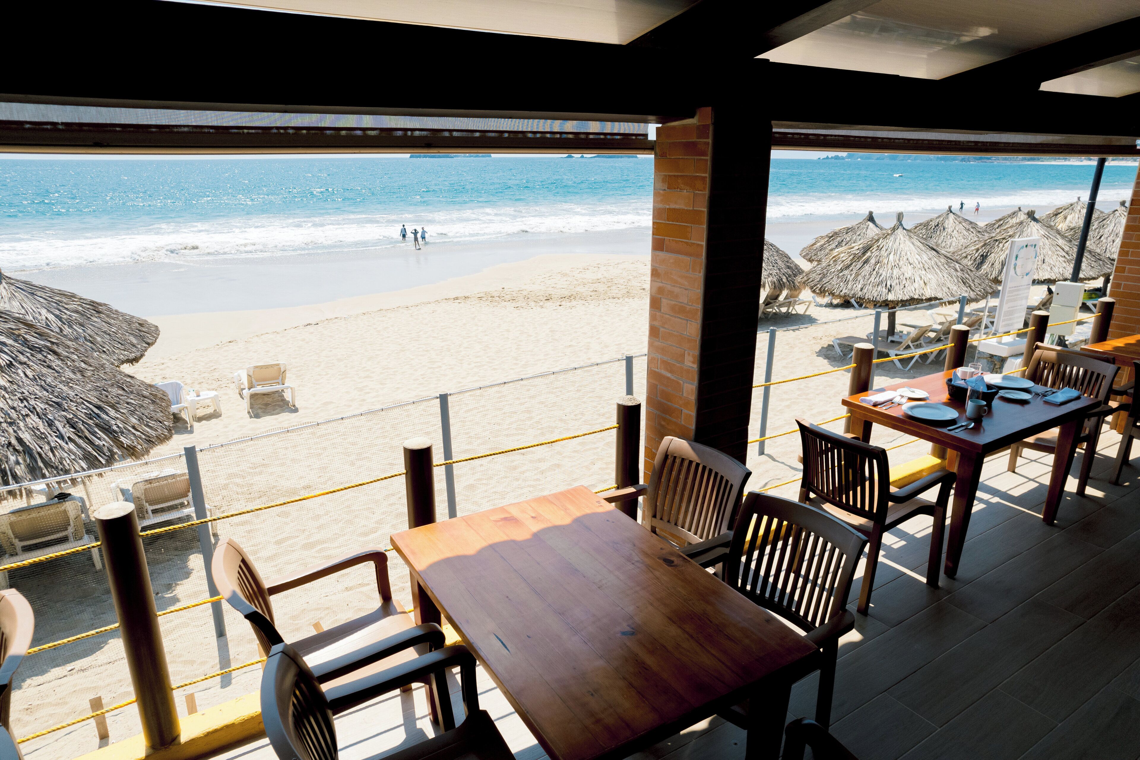 Un restaurante frente al mar con vista al océano, mesas, sillas y sombrillas de paja.