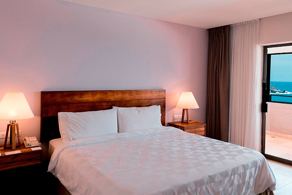 A hotel room with a large bed, two bedside lamps, a wooden headboard, and a view of the sea through a sliding door.