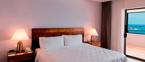 A hotel room with a large bed, two bedside lamps, a wooden headboard, and a view of the sea through a sliding door.