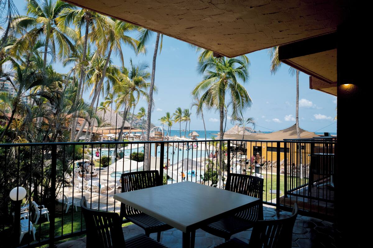A balcony with a table and chairs overlooking a pool area with palm trees and a view of the beach.
