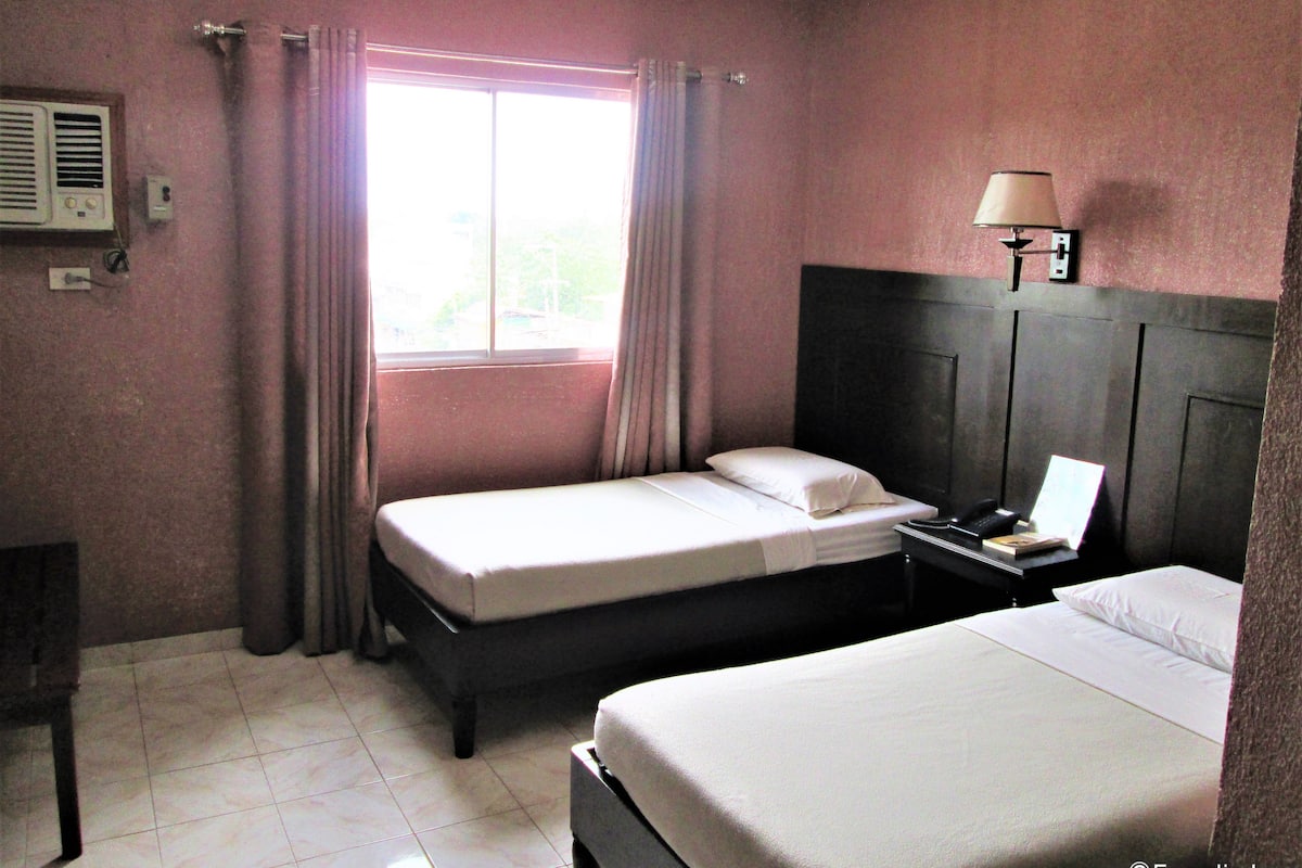 Habitación de hotel con dos camas, una ventana con cortinas, una mesita de noche con lámpara y un equipo de aire acondicionado instalado en la pared.