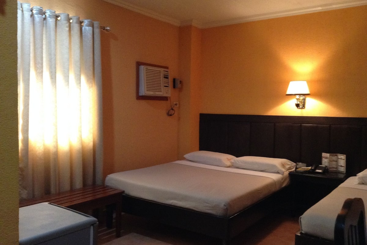 Habitación de hotel con una cama individual, una mesita de noche, una lámpara y una ventana con cortinas.
